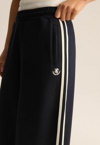 Granatowe dresy z białymi paskami po bokach, elastycznym pasem, kieszeniami bocznymi oraz małym haftowanym logo na lewej nodze. Miękka tekstura materiału.