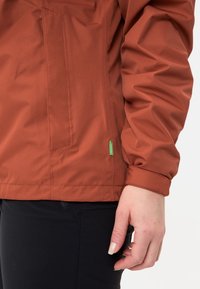 Rechterhand van een persoon in een roestkleurige jas met verstelbare manchet en zwarte broek, weergegeven van de taille tot halverwege het bovenbeen.