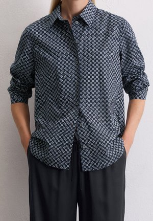 Osoba ubrana w koszulę z długim rękawem zapinaną na guziki z czarno-białym geometrycznym wzorem oraz czarne spodnie, z rękami w kieszeniach.