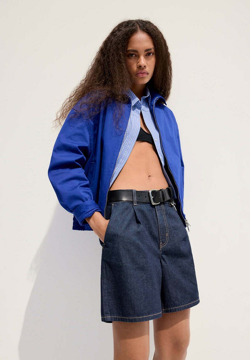 Woman with long curly hair wearing a blue jacket, partially open shirt, black belt, dark denim shorts, and black top standing against a white wall.