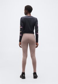 Άτομο που στέκεται με την πλάτη γυρισμένη φορώντας μαύρο μακρυμάνικο μπλουζάκι με πολύχρωμα σχέδια στα μανίκια και μπεζ κολάν με μαύρα αθλητικά παπούτσια.