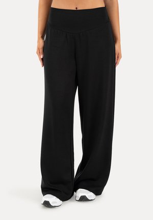 Mujer con pantalones negros anchos y sueltos y zapatillas blancas, de pie frente a un fondo claro y liso.