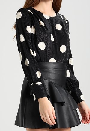 Vrouw draagt een zwarte blouse met grote witte polka dots, gecombineerd met een zwarte leren hoge taille rok met ruches.