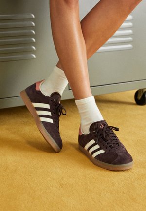 Osoba nosząca czarne buty sportowe Adidas z białymi paskami i różowym detalem na pięcie, białe skarpetki, stoi na żółtym dywanie blisko szarej szafki.