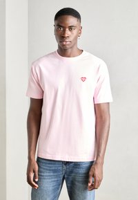 Lichtroze katoenen t-shirt met korte mouwen en een ronde hals, voorzien van een klein rood hartlogo op de linkerkant van de borst. Gemaakt in combinatie met een blauwe spijkerbroek.