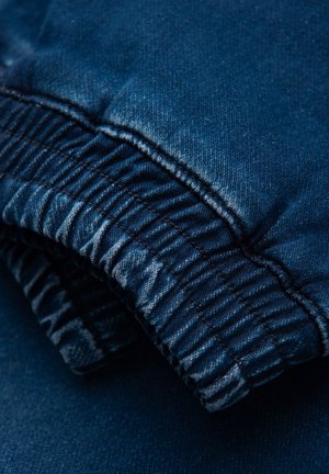 Ciemnoniebieski bawełniany denim, z elastycznym pasem w prążki i gładką teksturą. Zbliżenie ukazuje szczegóły szwów i subtelne wzory wyblaknięć.