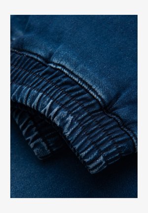 Ciemnoniebieski bawełniany denim, z elastycznym pasem w prążki i gładką teksturą. Zbliżenie ukazuje szczegóły szwów i subtelne wzory wyblaknięć.