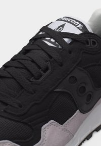 Κοντινό πλάνο μαύρου και γκρι αθλητικού παπουτσιού Saucony με υφασμένα κορδόνια, πάνελ από δίχτυ και σουέντ, και λογότυπο στη γλώσσα και στο πλάι.