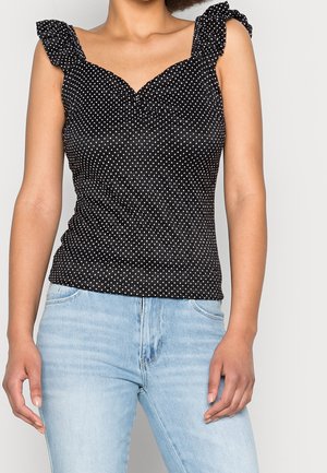 Vrouw draagt een zwart mouwloos topje met witte polkadots en gerimpelde bandjes, gecombineerd met lichtblauwe spijkerbroek, staand tegen een effen achtergrond.