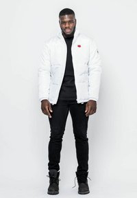 Vit puffjacka med hög krage, dragkedjestängning och en röd emblem, kombinerad med en svart t-shirt och svarta jeans. Svarta stövlar fulländar looken.