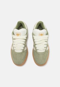 Las zapatillas de ante verde cuentan con acentos blancos, cordones texturizados y una suela de goma. El forro interior tiene un diseño estampado. La marca es visible en la lengüeta.