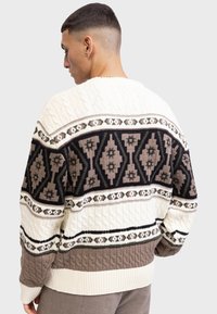 Mand iført en cremet, brun og sort mønstret kabelstrikket sweater, vendt for at vise ryg- og sideprofil mod en ensfarvet baggrund.