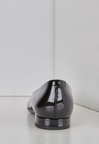 Černé lakové baleríny se hladkým, lesklým povrchem, kulatou špičkou a nízkým podpatkem. Viditelné švy po stranách podél zadní části.