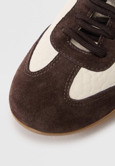 Καφετί σουέτ αθλητικό παπούτσι με λευκές υφασμάτινες λεπτομέρειες, με σκοτεινά κορδ όνια και στρογγυλεμένη μύτη. Μαλακό υλικό με αντίθετες ραφές.