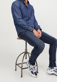Mörk denimskjorta med knapp-ned krage, i kombination med mörkblå skinny jeans och vita sneakers med blå detaljer, sittande på en metallpall.