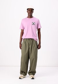 Lyserød bomulds t-shirt med en lomme grafik, parret med løstsiddende olivengrønne bukser. Modellen bærer en cremefarvet kasket og beige sneakers.