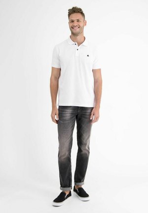 Lächelnder Mann, stehend, trägt ein weißes Poloshirt mit kurzen Ärmeln, ausgewaschene schwarze Jeans mit Umschlägen und schwarze Slip-On-Sneaker mit weißen Sohlen.