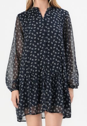 Frau trägt ein durchsichtiges marineblaues Kleid mit weißen Blumenmustern, langen Puffärmeln, Knopfleiste vorne und einem gerafften Saum.