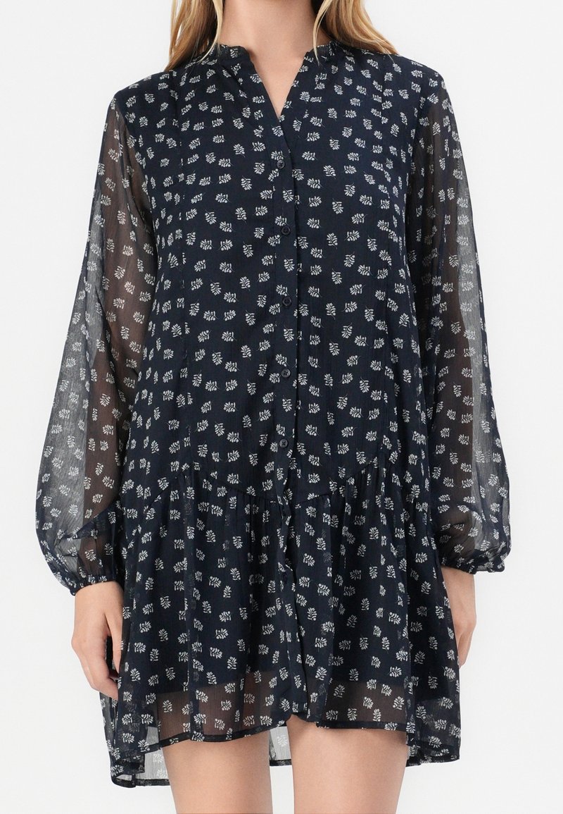 Femme portant une robe bleu marine transparente avec des motifs floraux blancs, longues manches bouffantes, devant boutonné et un ourlet volanté.