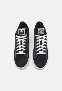 Sneakers in suede nero con lacci bianchi, caratterizzati da un design classico, cuciture a contrasto e un logo Adidas nero prominente sulla linguetta.