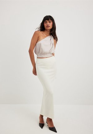 Femme debout devant un fond uni, portant un haut beige à une épaule, une longue jupe blanche et des talons noirs pointus, regardant vers la caméra.