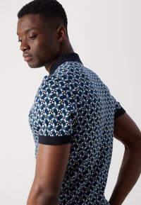 Granatowa koszulka polo z geometrycznym wzorem w białym i jasnoniebieskim kolorze. Prążkowany kołnierzyk i mankiety z teksturowanym wykończeniem materiału.