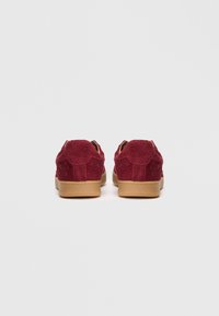 Røde teksturerte joggesko med et mykt stoffoverdel, rund tå og beige gummisåle. Har minimalistisk design og ingen synlig maskinvare.