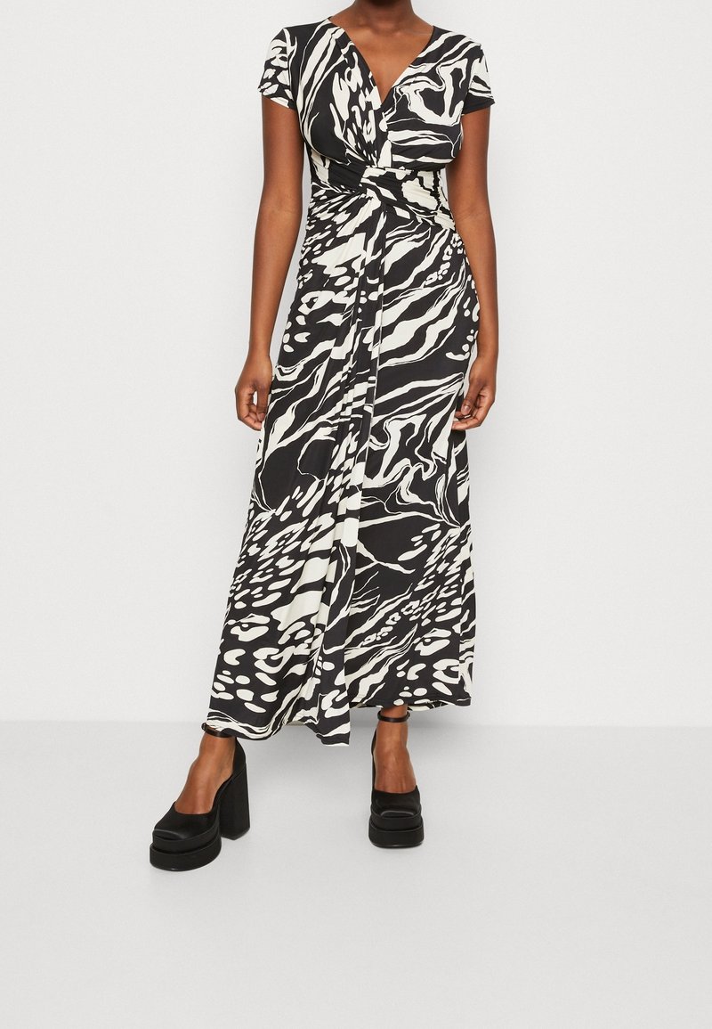 Femme portant une robe portefeuille à motifs abstraits noir et blanc, à manches courtes, avec des talons compensés noirs.
