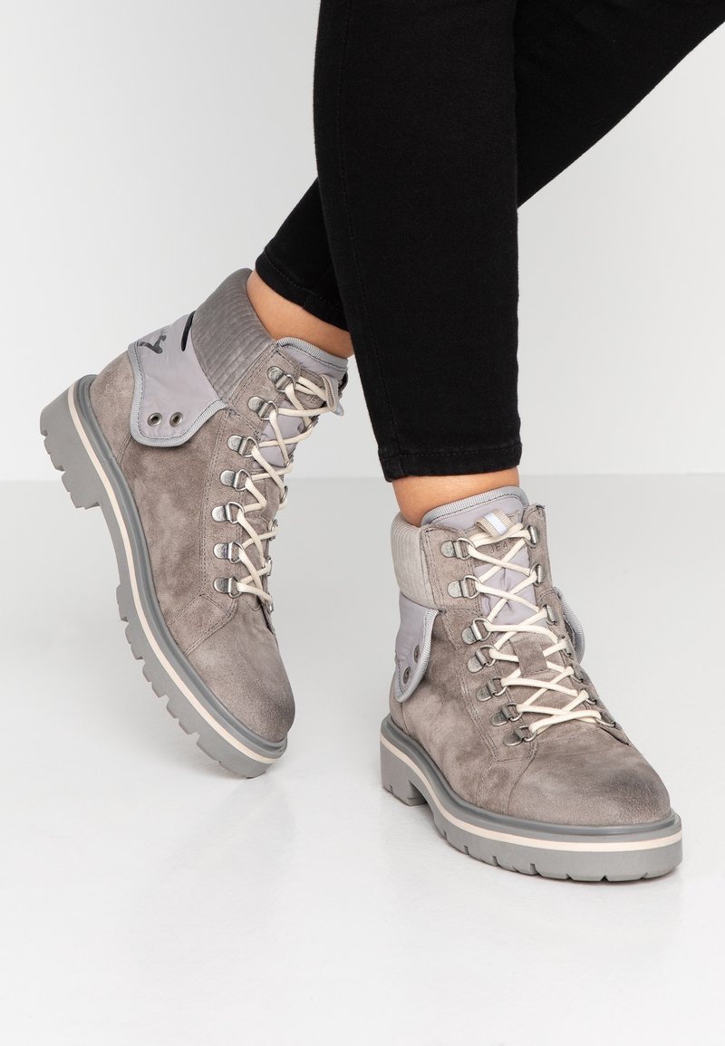 Persona que lleva botas de senderismo de ante gris con tobillos acolchados y cordones blancos, combinadas con pantalones negros ajustados sobre un fondo liso.