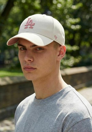 Young man wearing a light pink Los Angeles cap and gray t-shirt stands outdoors with blurred greenery and stone wall in the background.
