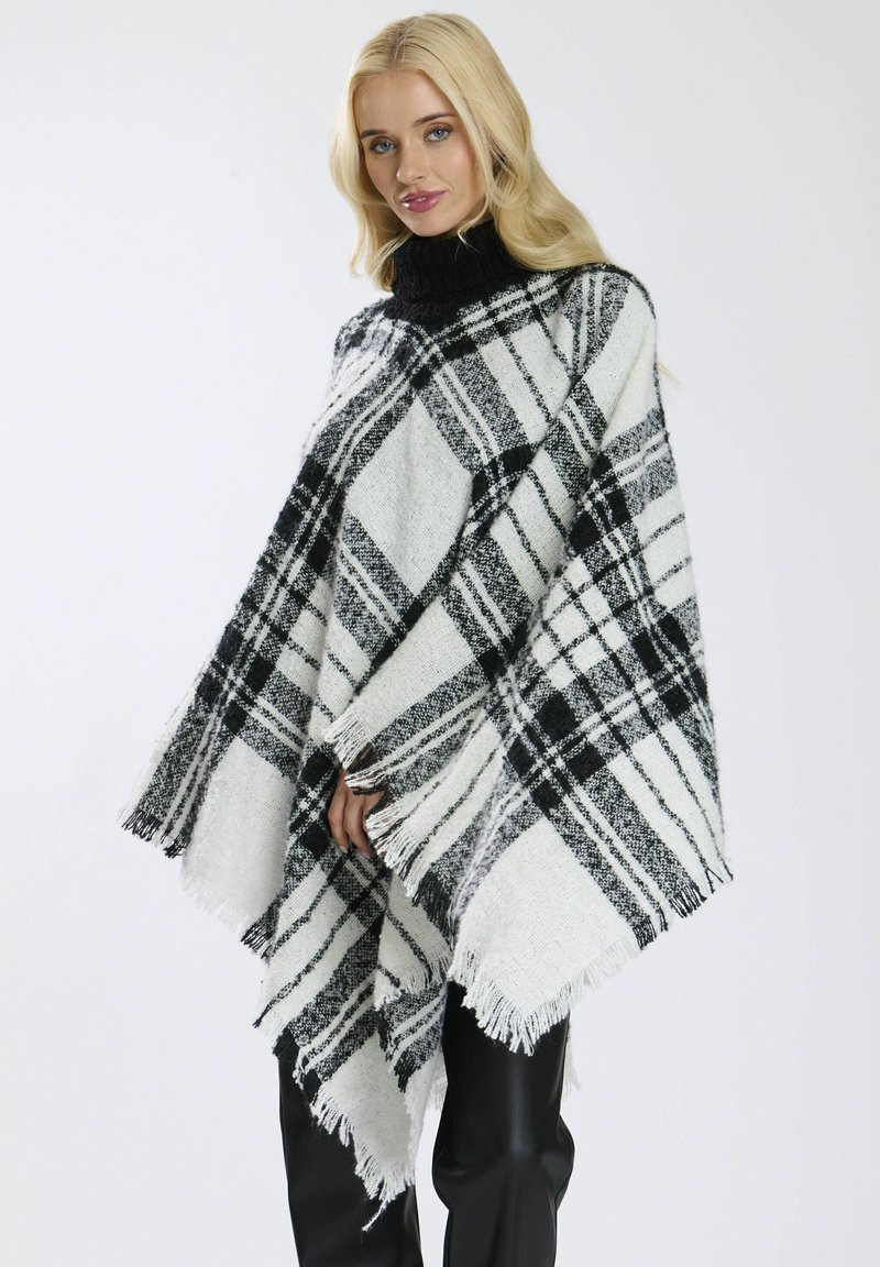 Poncho à carreaux noir et blanc avec un ourlet à franges et un col montant en tricot noir, fabriqué en tissu texturé. Porté par-dessus un pantalon noir.