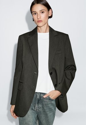 Blazer grigio scuro su misura con rever a punta, chiusura a un bottone, tasche frontali, indossato sopra una t-shirt bianca e jeans blu.