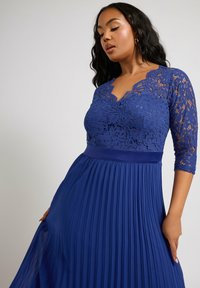 Blå spetsklänning med V-ringning och trekvartsärmar. Plisserad kjol ger struktur, med ett fast midjeband och blomster-spetsdetaljer.