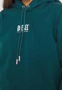 Turkusowa bluza z kapturem, ze sznurkiem do regulacji, z białym logo na piersi. Gładka faktura i luźny krój, z metalowymi końcówkami na sznurku.