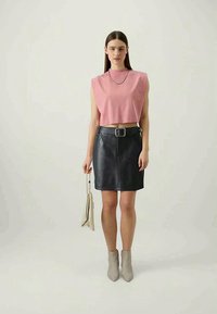 Fekete bőr szoknya elöl zippzárral és övvel, rózsaszín, ujjatlan, cropped felsővel és bézs bokacsizmával párosítva, egy kis táskával a kezében.