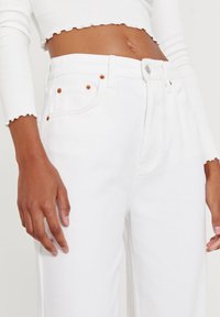 Zbliżenie na osobę ubraną w białe spodnie z wysokim stanem i białą krótką bluzkę z długim rękawem o falbaniastych krawędziach.