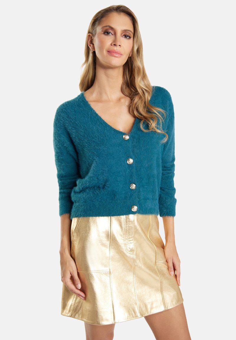 Cardigan en peluche de couleur bleu canard avec un décolleté en V profond et cinq boutons dorés brillants. Assorti à une mini-jupe métallique dorée avec des détails de couture.