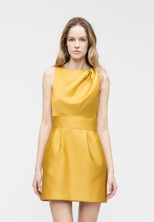 Femme aux longs cheveux blonds portant une robe en satin jaune moutarde sans manches avec un détail froncé près de l'encolure, debout devant un fond uni.