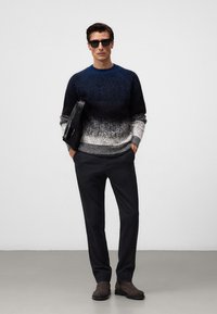 Sweter dzianinowy z gradientowym wzorem od granatowego do szarego, połączony z ciemnymi spodniami i brązowymi skórzanymi butami. Modelka trzyma czarną torbę.