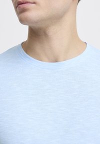 Světle modré tričko s krátkým rukávem a kulatým výstřihem, vyrobené z měkké, texturované látky. Má hladký límec a jednoduchý, minimalistický design.