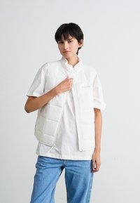Persoon met kort zwart haar in een witte gewatteerde vest over een wit t-shirt en een blauwe spijkerbroek, staand tegen een effen lichte achtergrond.