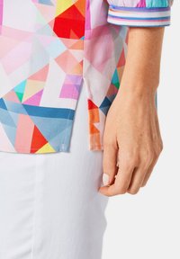 Camicia colorata con un motivo geometrico nei toni del rosa, blu e rosso, con un dettaglio di pattina e un polsino a strisce. Tessuto bianco sotto.