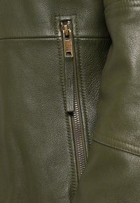 Olijfgroene leren jas met een getextureerd oppervlak, voorzien van een metalen ritssluiting en een merkgebonden ritstrekker.