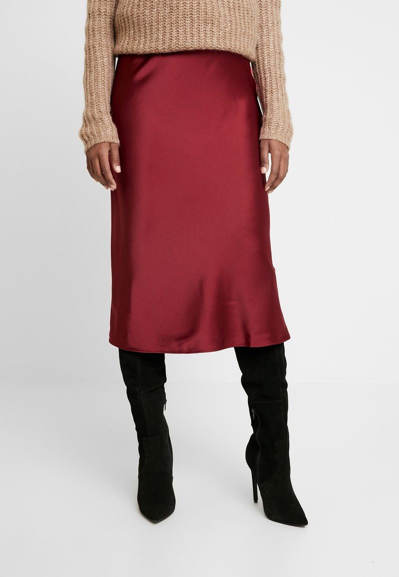 Falda midi de satén carmesí con una textura suave y un ligero brillo. Combinada con botas de tacón alto negras hasta el muslo y un suéter de punto beige.