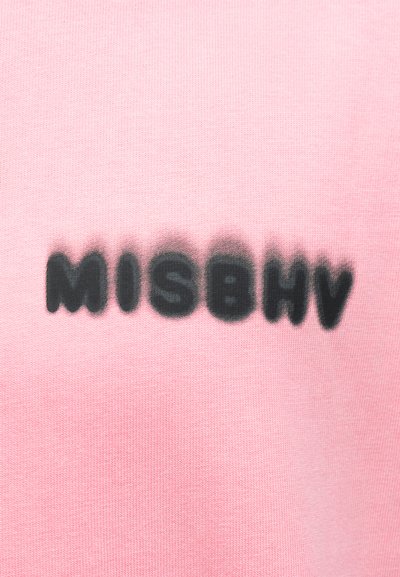 Η λέξη «MISBHV» ψεκασμένη με μαύρο σπρέι σε ροζ υφασμάτινη επιφάνεια, με θολό εφέ σκιάς γύρω από τα γράμματα.