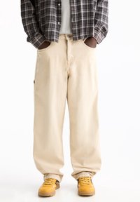 Personne portant un pantalon beige à jambes larges, des baskets jaunes et une chemise à carreaux noir et blanc, les mains dans les poches.