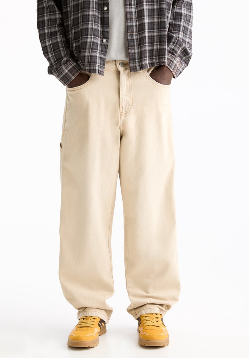 Personne portant un pantalon beige à jambes larges, des baskets jaunes et une chemise à carreaux noir et blanc, les mains dans les poches.