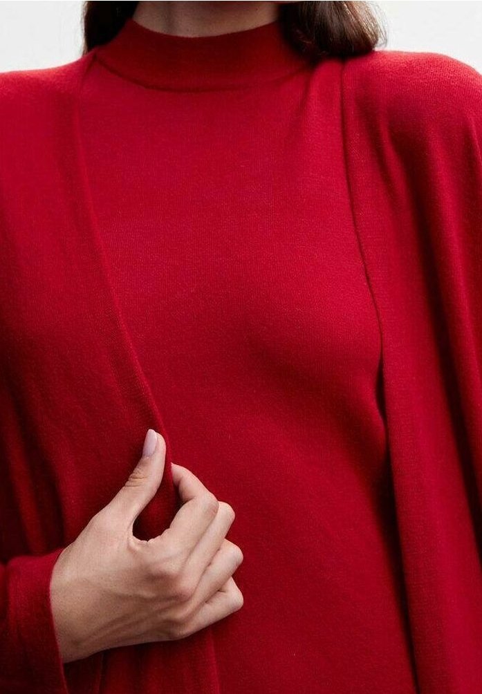Femme portant un pull à col roulé rouge tenant le bord d'un cardigan assorti rouge sur un fond uni.