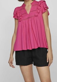 Rosa bomullsblus med volangärmar och V-ringning, med rynkade detaljer. Matchad med svarta strukturerade shorts.