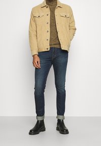 Homme portant une veste en velours côtelé beige, un pull en laine marron, un jean bleu foncé retroussé et des bottes en cuir noir, debout devant un fond blanc.